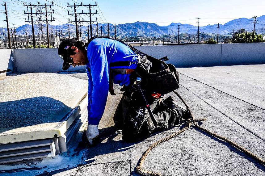 Man in blue shirt with black toolbelt working on a skylight that can cause roof leaks on an industrial roof with telephone poles and mountains in the background