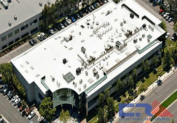 Arial view of white industrial building with white roof with skylights and Air Conditioning units surrounded by green trees